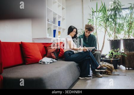 Two students studying together on a couch in a bright indoor lounge with plants and backpacks Stock Photo