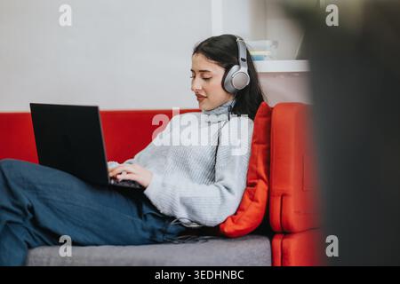 Young student using laptop and headphones on a red couch studying remotely at home Stock Photo