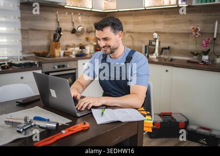 Handyman wearing blue uniform using laptop in kitchen with plumbing tools on the table Stock Photo