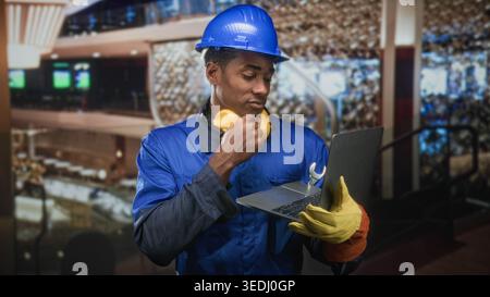 Man in hardhat wearing blue coverall and noise cancel headphones holding laptop with wrench and hand to chin in building; focused problem solving. Stock Photo
