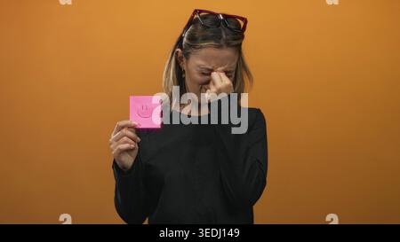 Young blonde woman pinches nose with thumb and forefinger while holding pink sticky note with smile drawing against orange studio wall, wearing black Stock Photo