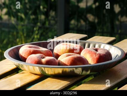 Fresh flat peaches in a metal bowl on a wooden outdoor table, captured in soft natural light for a fresh seasonal look. Stock Photo