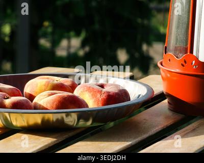 Flat peaches in a metal bowl and red lantern on a wooden table, creating a seasonal fruit still life with a rustic natural setting Stock Photo