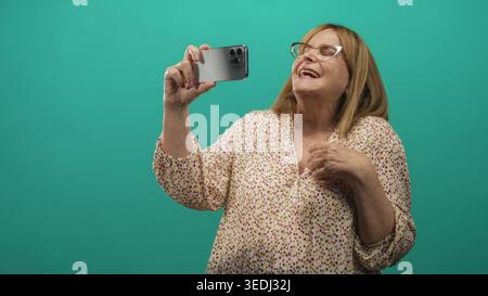 Senior woman wearing glasses and floral blouse holds smartphone with raised arm and hand on chest in studio; joy connection. Stock Photo