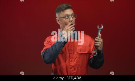 Young mechanic man in orange coverall holds a wrench and places hand to mouth gesture, wearing glasses against a red studio backdrop while inspecting Stock Photo