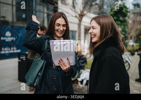 Two female students laughing and studying together outdoors in an urban campus setting Stock Photo