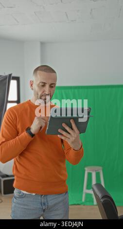Man holding tablet and gesturing with hand in studio among softbox lights and green screen, wearing orange sweater and jeans; creative concentration. Stock Photo