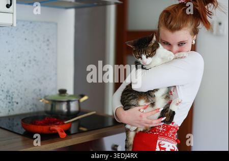 Cozy kitchen scene with young woman, homemade cooking and affectionate cat in arms. Stock Photo