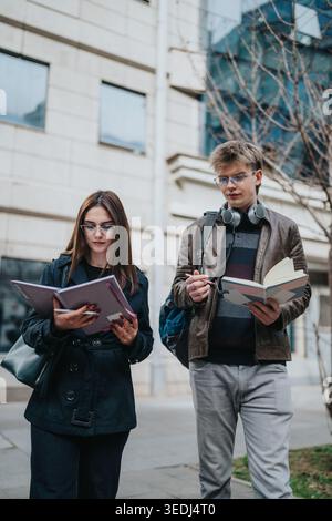 Two students learning outdoors reading notebooks while walking near university buildings Stock Photo