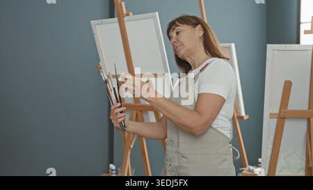 Woman painter selects paintbrushes and adjusts brush tips before blank canvas in studio; creative focus. Stock Photo