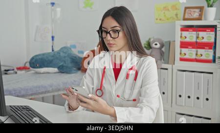Young woman doctor with stethoscope holding smartphone, hands visible and scrolling while sitting at computer desk in a building; quiet concentration. Stock Photo