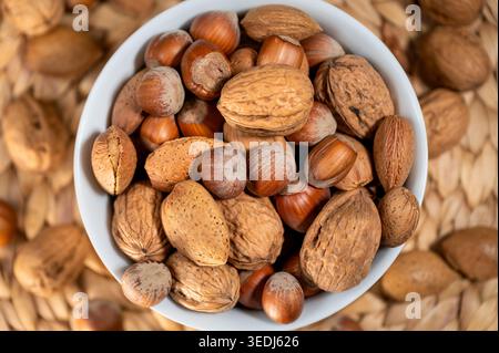 Assortment of whole nuts in shell including walnuts, almonds and hazelnuts in a white bowl on a wicker surface, shot from above. Stock Photo