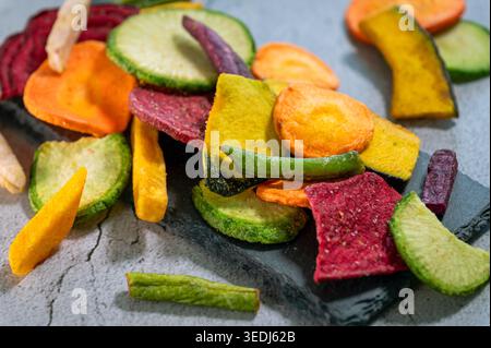 Assortment of colorful dehydrated vegetable chips including beetroot, carrot, zucchini and sweet potato scattered on a slate and stone surface. Stock Photo