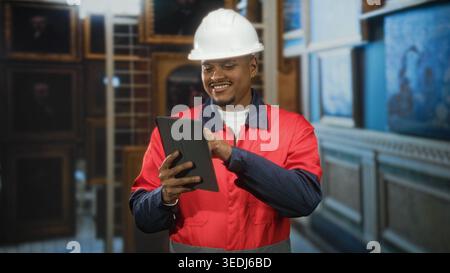 Man in hard hat and red uniform tapping a tablet with finger inside a building, pointing gesture during inspection of gallery paintings; confidence. Stock Photo
