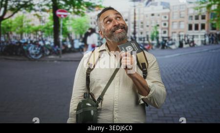 Man holding passport and gripping backpack strap on city street, smiling and looking upward; travel joy. Stock Photo