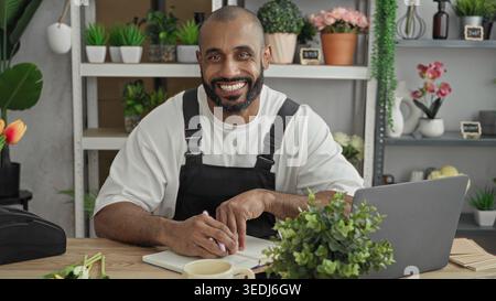 Man florist in apron writes in notebook by laptop at studio desk, pen in hand; calm business planning. Stock Photo