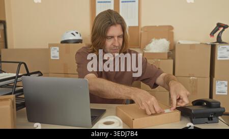 Man with blond hair writing a shipping label on a cardboard parcel with his hands at a cluttered desk with laptop, tape roll and stacked boxes in a pa Stock Photo