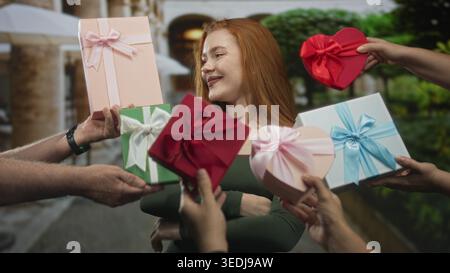 Redhead woman smiling and receiving wrapped gift boxes from multiple hands on a street, hands offering heart box and ribboned parcels; joy celebration Stock Photo