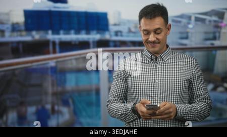Man with moustache wearing checkered shirt holding smartphone, fingers typing a message while standing at an airport terminal railing; contentment. Stock Photo