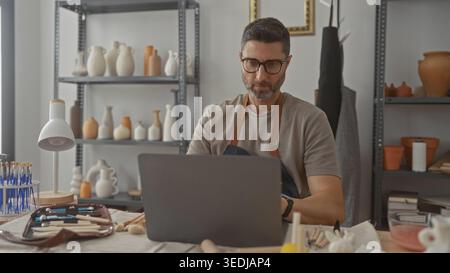 Man potter with hand on chin using laptop in studio among pottery shelves, brushes and clay tools; thoughtful craft practice. Stock Photo