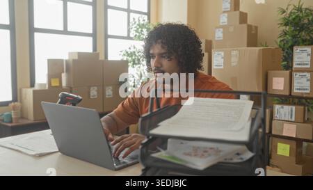 Man in orange shirt holding document and hand on cheek beside laptop and barcode scanner in building; small business concentration. Stock Photo