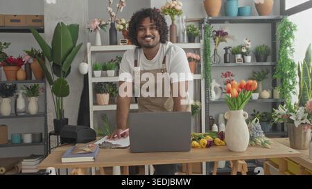 Man florist in apron leans on table with laptop and tulips, hands writing on clipboard in studio; warmth and craft. Stock Photo