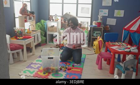 Man kneels assembling and sorting toy blocks into a clear bin on foam number mat inside a kindergarten building; patience nurture. Stock Photo
