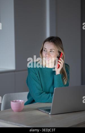 Contented middle-aged woman sitting at table with laptop, talking on phone while working from home. Calm focused expression, modern lifestyle of remot Stock Photo