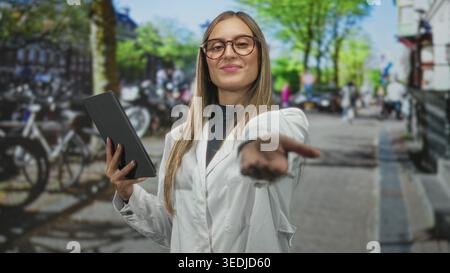 Woman holding tablet reaches out on a busy city street with bicycles and parked cars, wearing white coat and glasses; digital confidence. Stock Photo