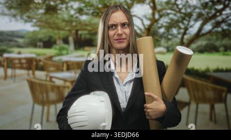 Woman holding white hardhat and rolled blueprint tubes smiling on an outdoor building terrace while wearing blazer and clutching plans; confidence car Stock Photo