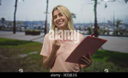 Young woman holding a red clipboard reading and smiling on street by marina promenade with palm trees and boats visible; calm focus optimism. Stock Photo