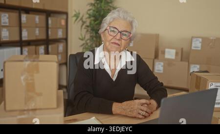 Elderly woman smiling with hands clasped by laptop and packing box in a building small business shipping station, surrounded by stacked parcel boxes; Stock Photo
