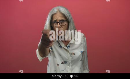 Middle aged grey haired woman pointing finger in a studio with pink backdrop; anger accusation demand. Stock Photo