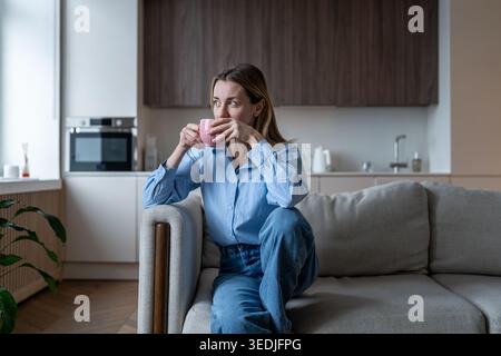 Calm midlife woman enjoys aroma coffee break on sofa dreamy looking to window taking mindful break. Stock Photo