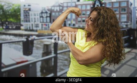 Woman flexes bicep and points to arm on a canal street beside an iron bridge and waterfront walkway, wearing a yellow top and smiling; pride strength Stock Photo