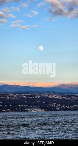 A vertical landscape featuring a bright full moon in a soft blue sky above a sprawling hillside city. Pink-tinged clouds rest on distant mountain peak Stock Photo