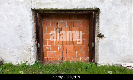 A sealed entrance featuring a dark timber frame filled with modern orange bricks. The surrounding facade has cracked plaster and a base of green grass Stock Photo