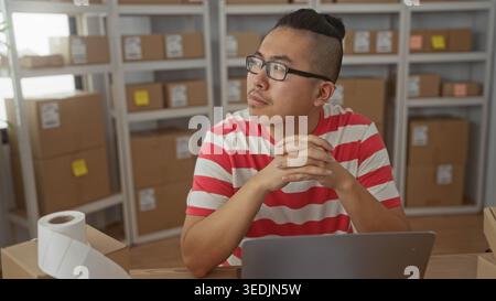 Man clasping hands over laptop and parcel at a desk with cardboard boxes on shelves in a building, wearing glasses and striped shirt; thoughtful plann Stock Photo