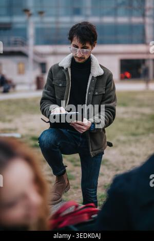 Young high school student using a tablet outdoors on an urban campus park bench in jacket Stock Photo