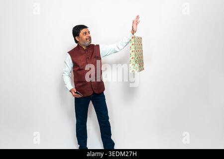 A man wearing a traditional vest and jeans is shown holding a shopping bag in one hand and a smartphone in the other against a plain background. Stock Photo