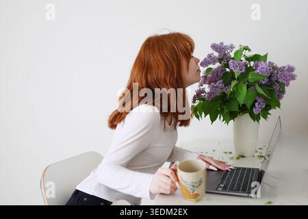Young redhead woman smelling lilac flowers while working on laptop at home, creating cozy spring lifestyle and remote work atmosphere. Stock Photo