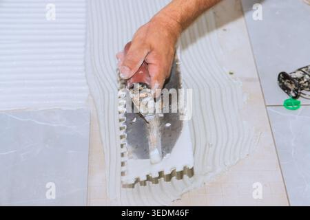 Tiler worker uses trowel to spread adhesive on floor surface during bathroom renovation task at home. Stock Photo