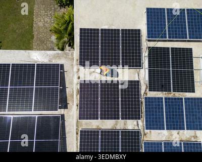 Aerial view of a technician installing solar panels on a rooftop with lush greenery nearby in Guanaja, Bay Islands Department, Honduras. Stock Photo