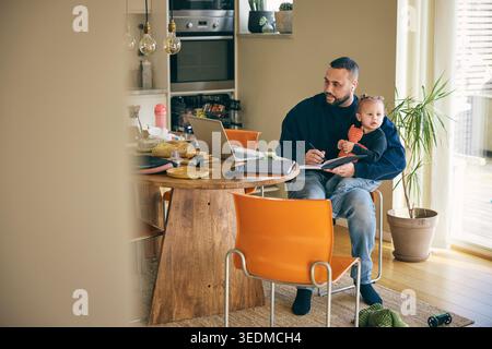 Focused man writing notes through laptop while sitting with daughter at table in home office Stock Photo