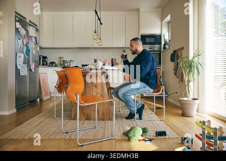 Male freelancer drinking coffee while working on laptop from home Stock Photo