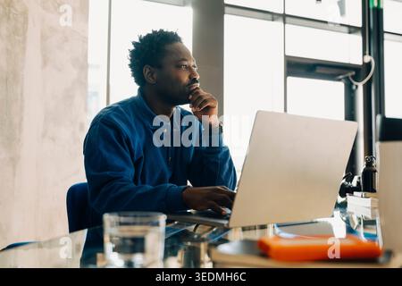 Thoughtful male business professional sitting with laptop at desk in modern office Stock Photo