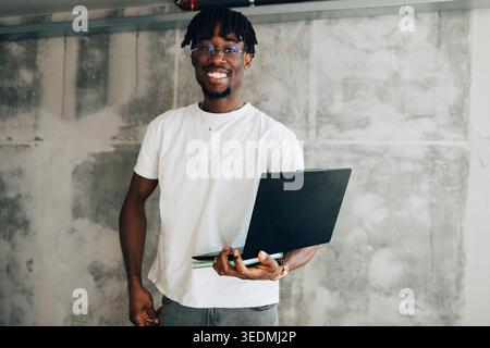 Portrait of confident businessman holding laptop while standing against wall in office Stock Photo
