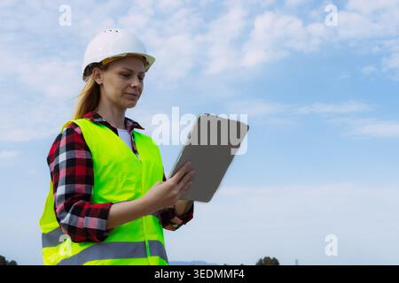 Woman engineer, dressed in safety gear, actively managing and inspecting site operations with a digital tablet, symbolizing technological integration Stock Photo