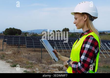 Female engineer wearing a helmet and safety vest, checking readings on a digital tablet at a large solar panel farm, monitoring renewable energy produ Stock Photo