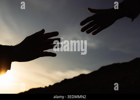 Two silhouetted hands reaching towards each other in a gesture of support during an inspiring sunset, conveying concepts of rescue, collaboration, par Stock Photo